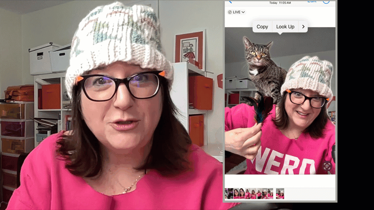 A woman in a pink sweater and knitted hat holds a phone showing an image of her with a cat on her head. She appears to be on a live video call, perhaps demonstrating how to create an Apple Live Sticker.